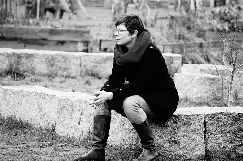 Frau im schwarzen Mantel sitzt auf einem Stein und schaut nach links