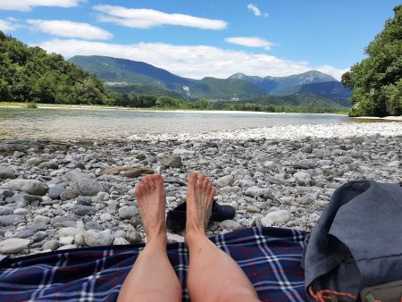 Füße auf einer Decke im Flussbett des Tagliamentos. Dahinter Berge.